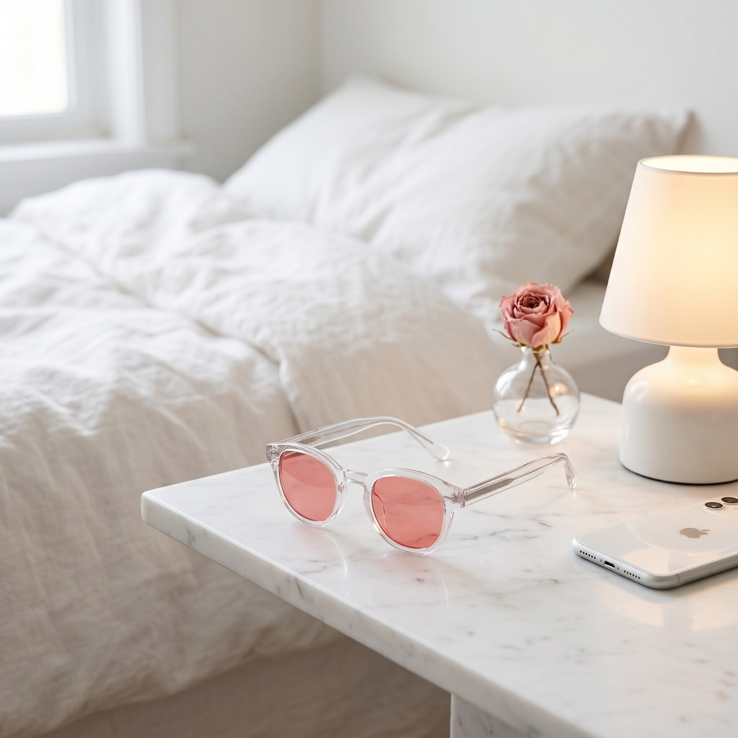 Pink-lensed sunglasses, a smartphone, and a rose in a vase rest on a marble bedside table.