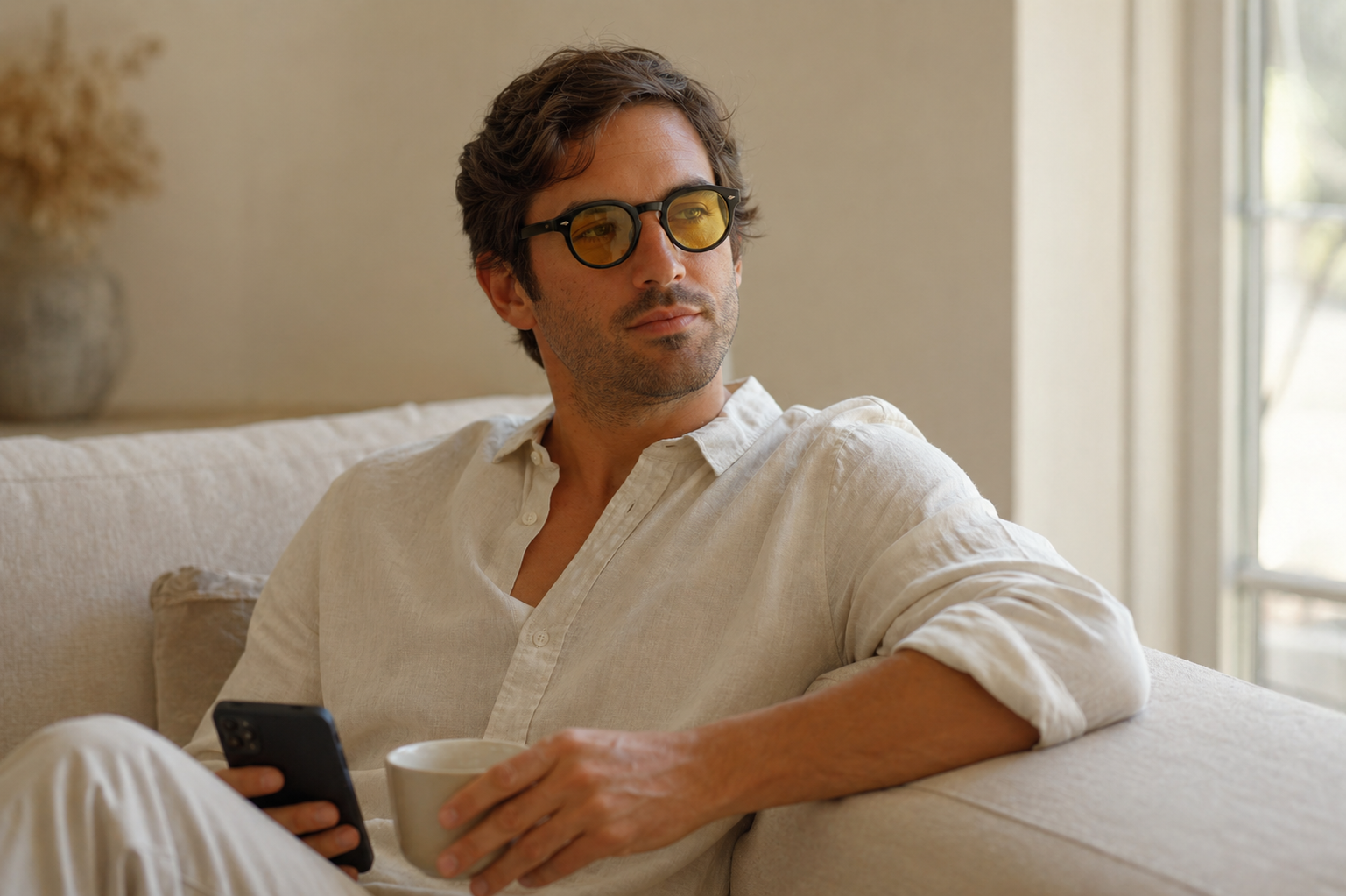 A man in yellow-tinted glasses sits on a couch, holding a phone and a mug, looking away.