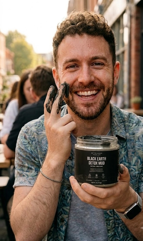 A smiling man holds a jar of Black Earth Detox Mud while applying it to his cheek.