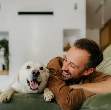 A smiling man with a beard lies on a green couch next to a happy white dog.