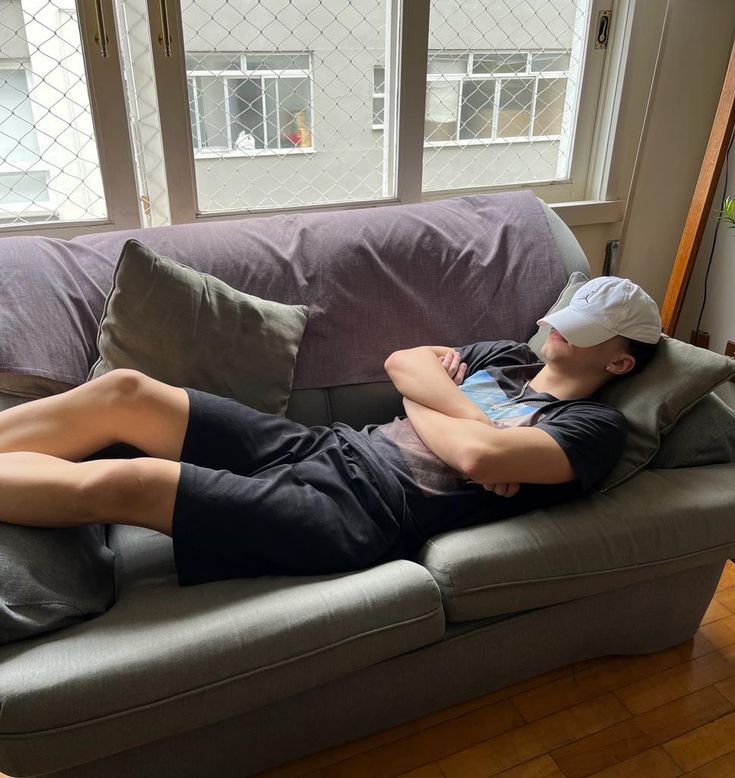 A young man wearing a white baseball cap is sleeping on a grey couch with his arms crossed.