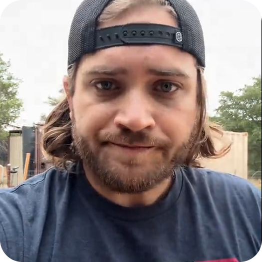 Man with beard wearing a cap and blue t-shirt outdoors.