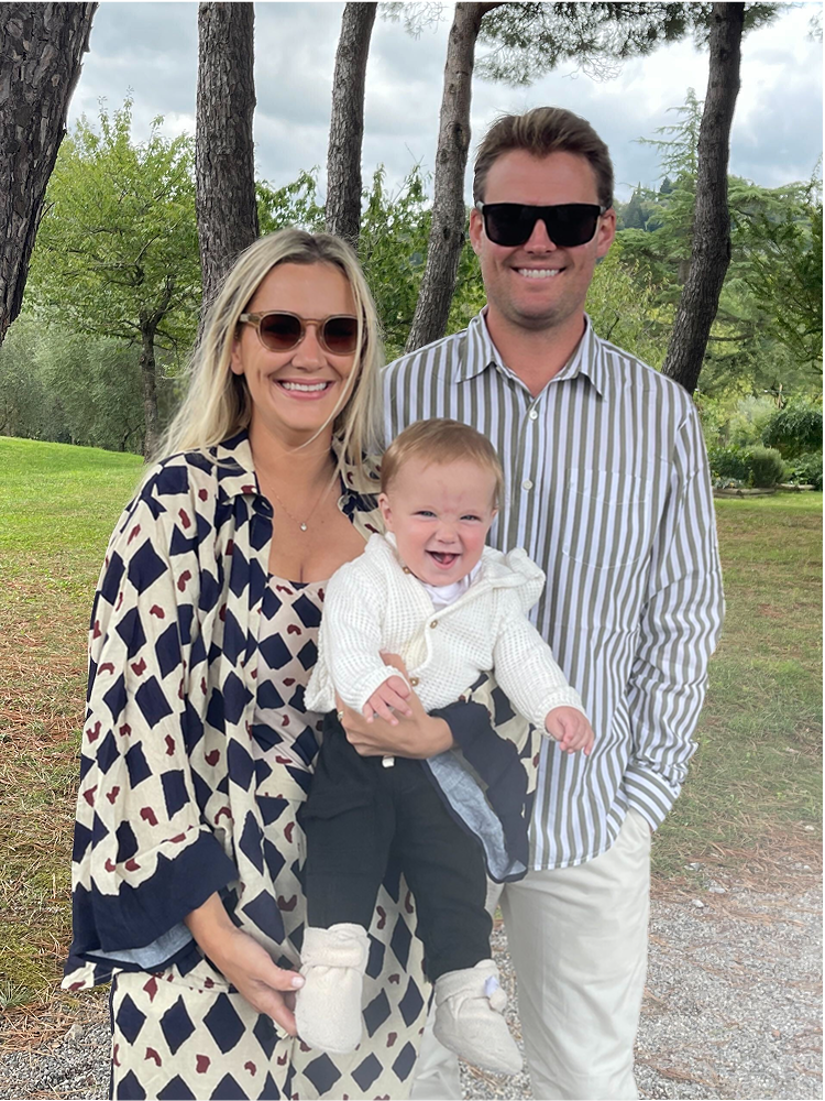 Smiling couple with a baby in an outdoor setting with trees and grass.
