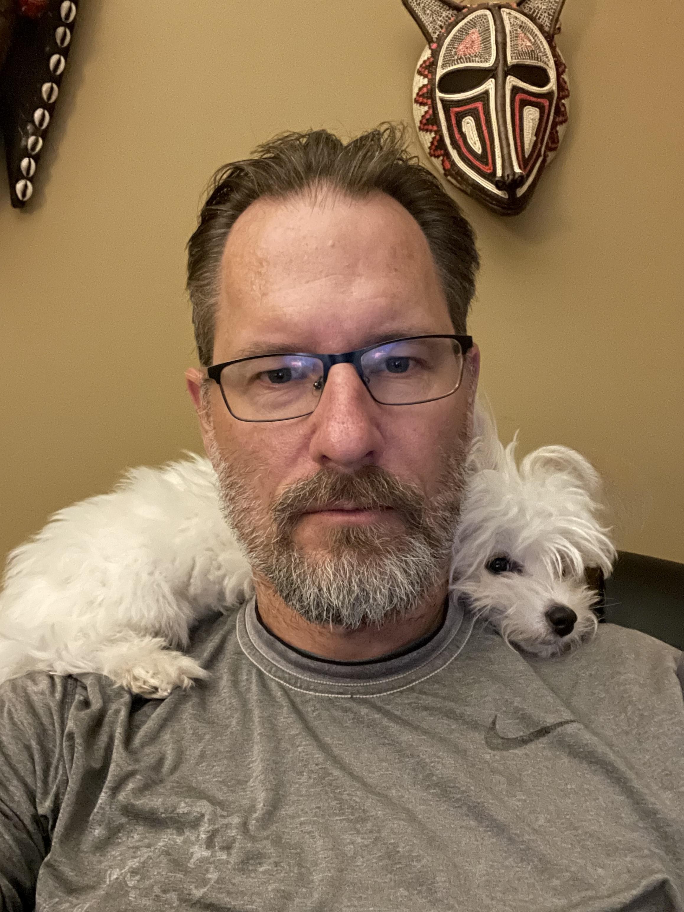 Man with glasses and a small white dog on his shoulder.