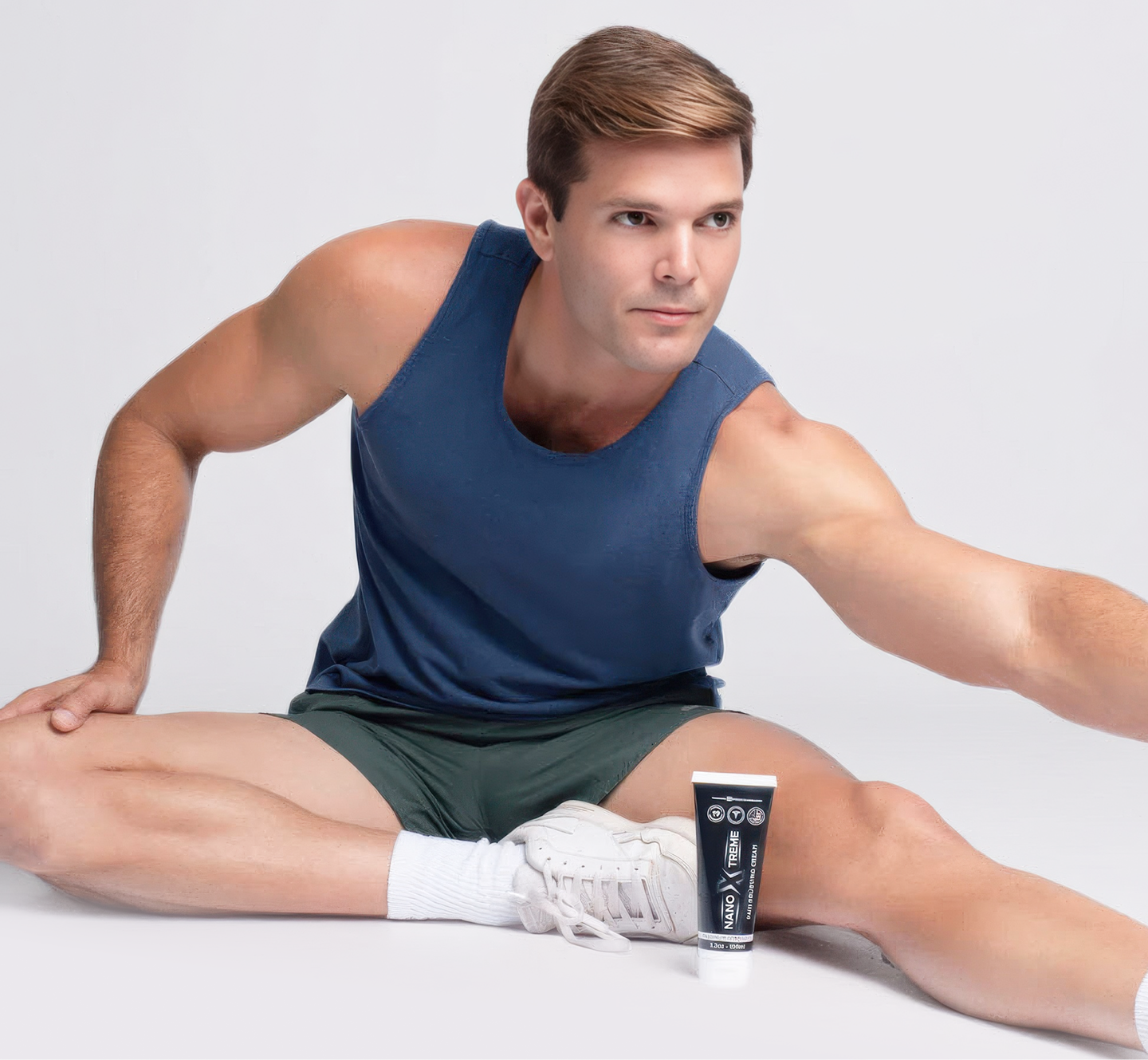 A man in a blue tank top stretches on a white floor with a tube of cream by his foot.