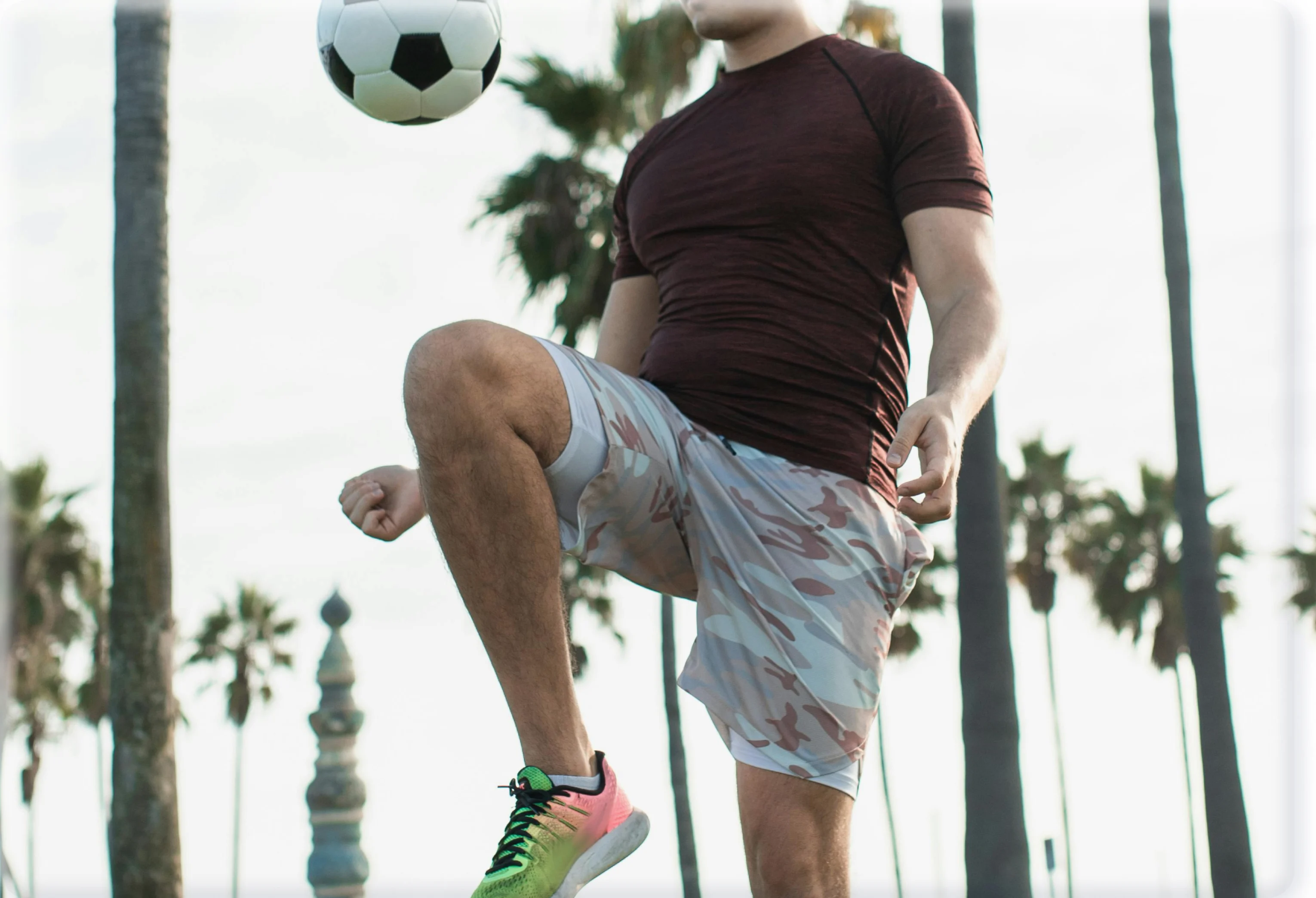 A man in athletic wear juggles a soccer ball on his knee, with palm trees in the background.