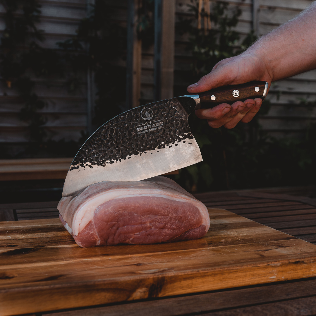 A hand holding a cleaver above a piece of raw meat on a wooden board.