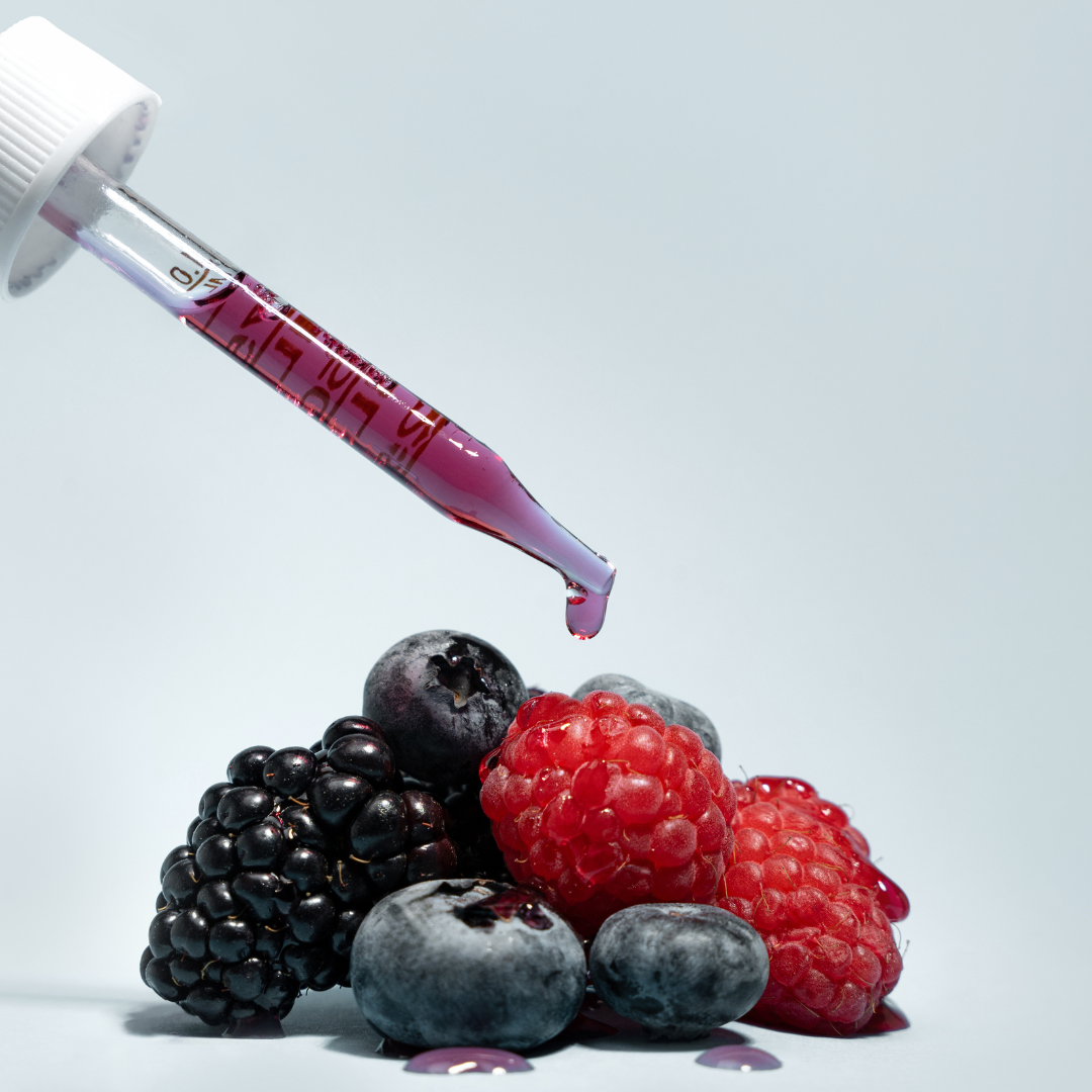 Glass dropper dispensing a pink berry-flavored liquid supplement onto fresh raspberries and blueberries.