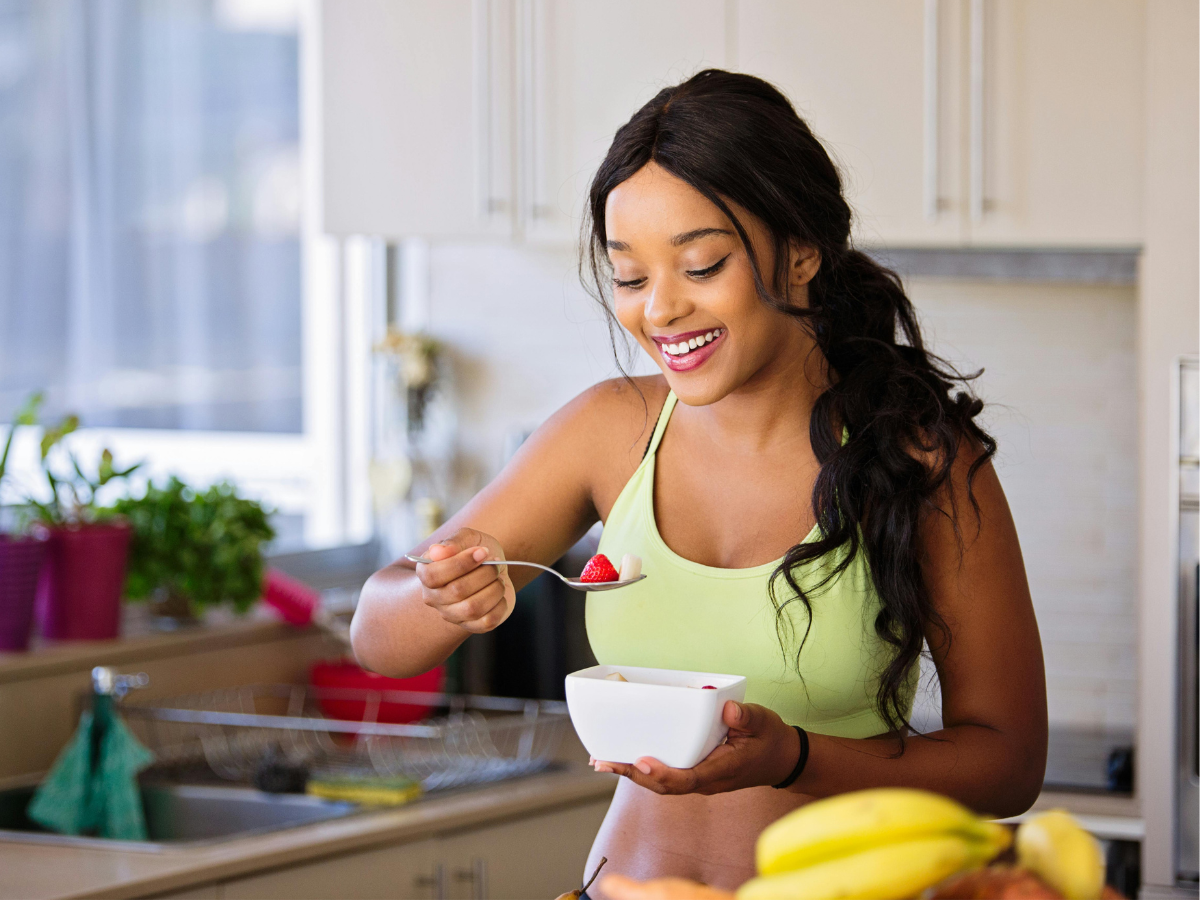 A smiling woman in a lime green sports top eats fruit from a white bowl in a kitchen.