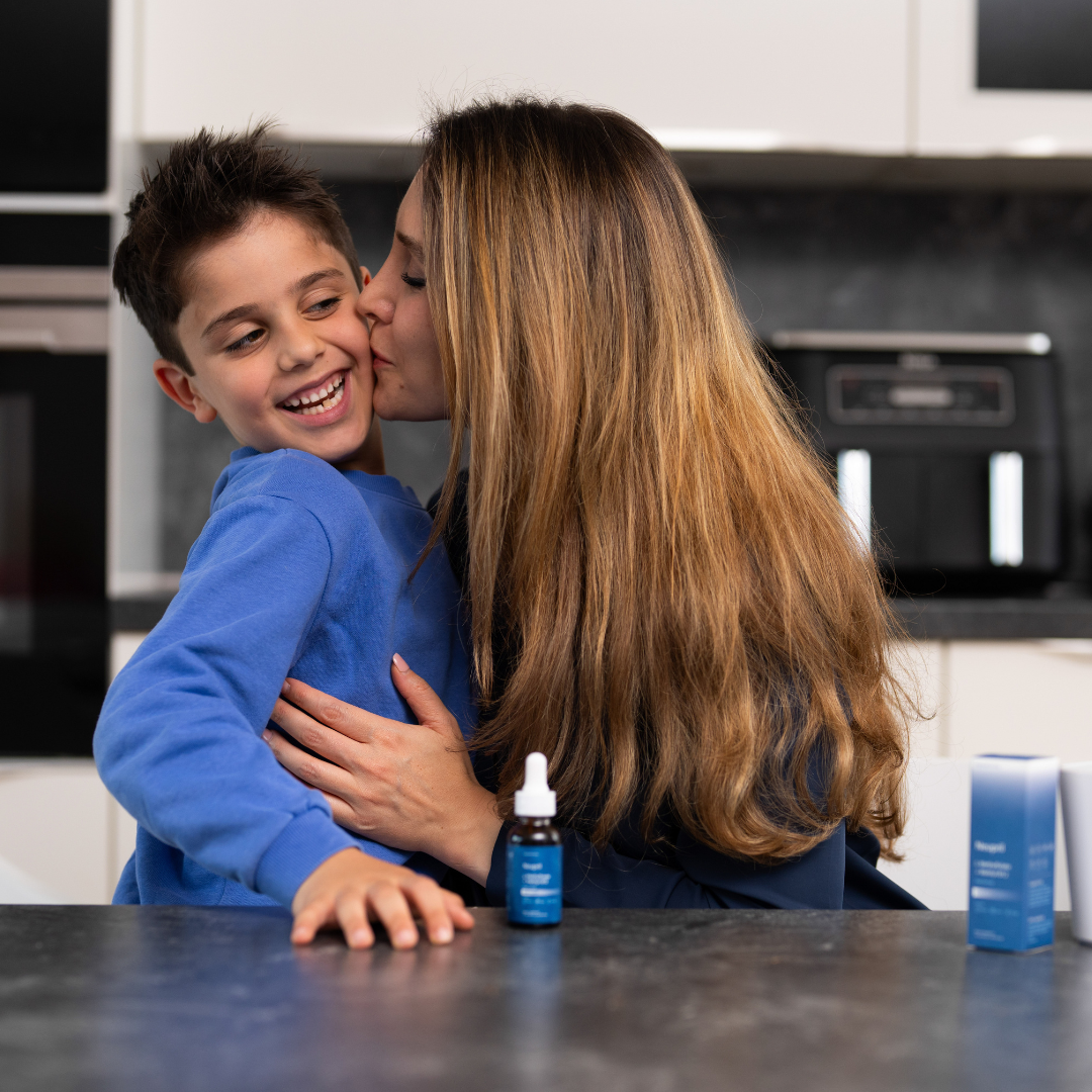 A mother kisses her smiling son’s cheek as he hugs her at a kitchen counter. In the foreground are a bottle of Neupril Kids L-Methylfolate + Methyl-B12 liquid supplement and its product box, with a modern kitchen in the background.