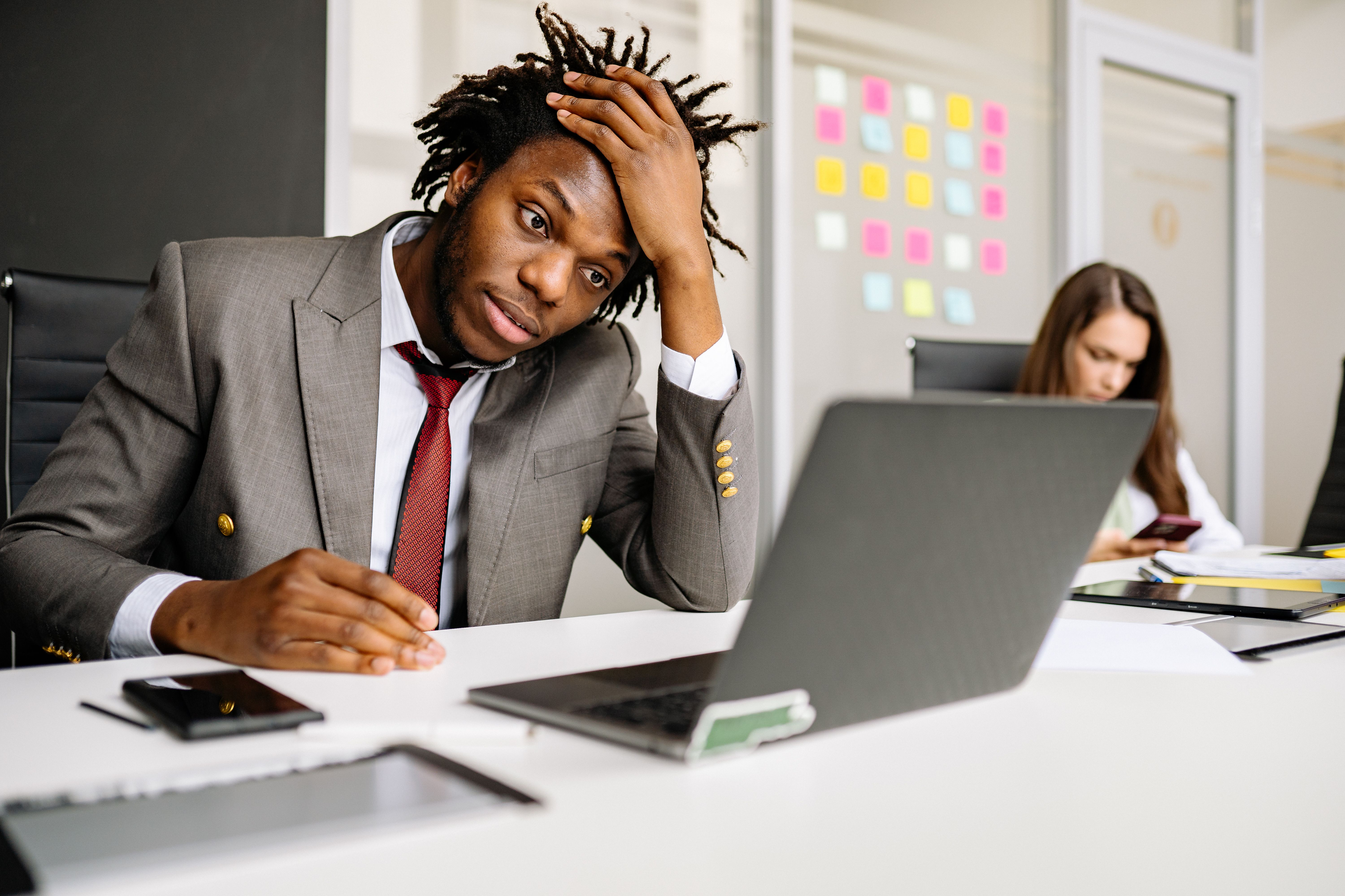 A young Black man wearing a grey business suit and red tie sits at a white office desk, looking overwhelmed and holding his hand to his head while staring intensely at a laptop screen.