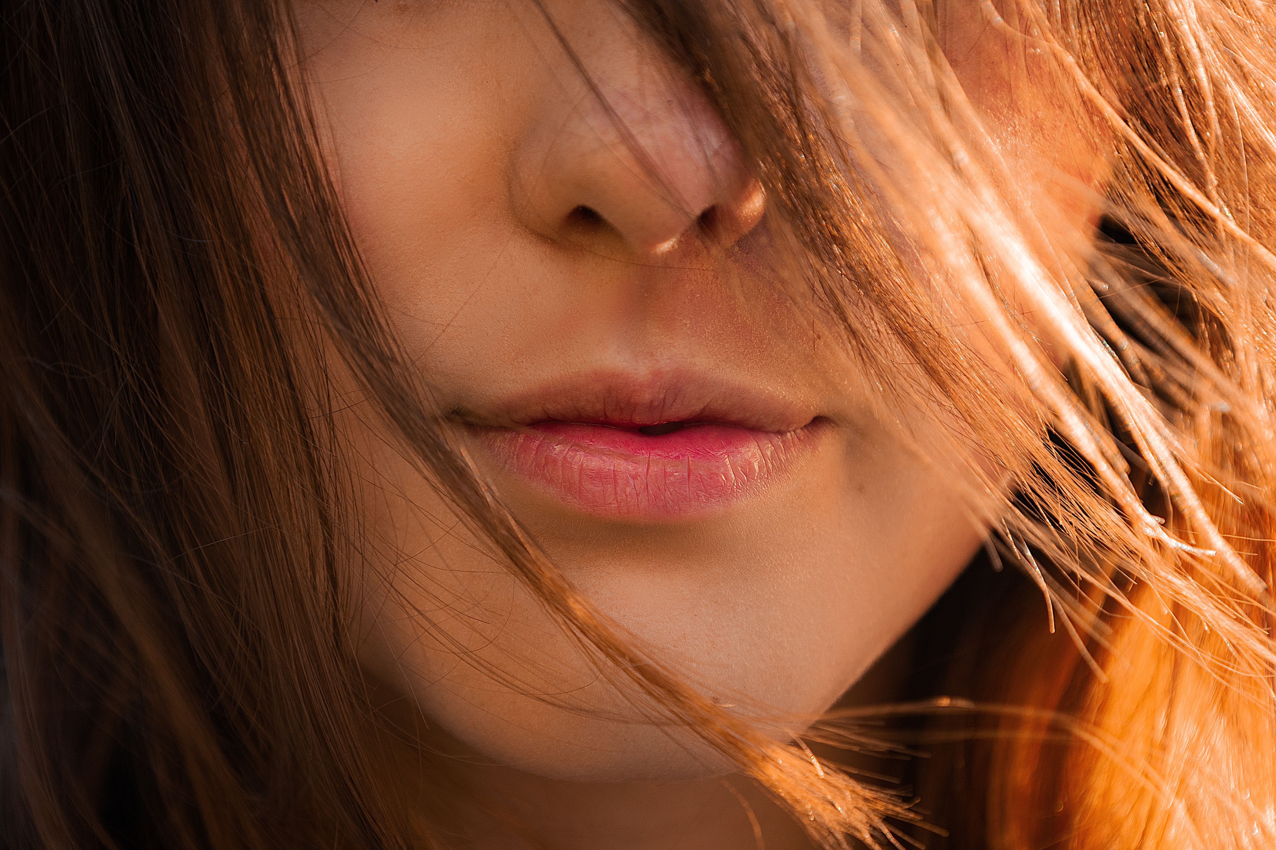 A close-up of a person's mouth and nose, with their sunlit hair blowing across their face.
