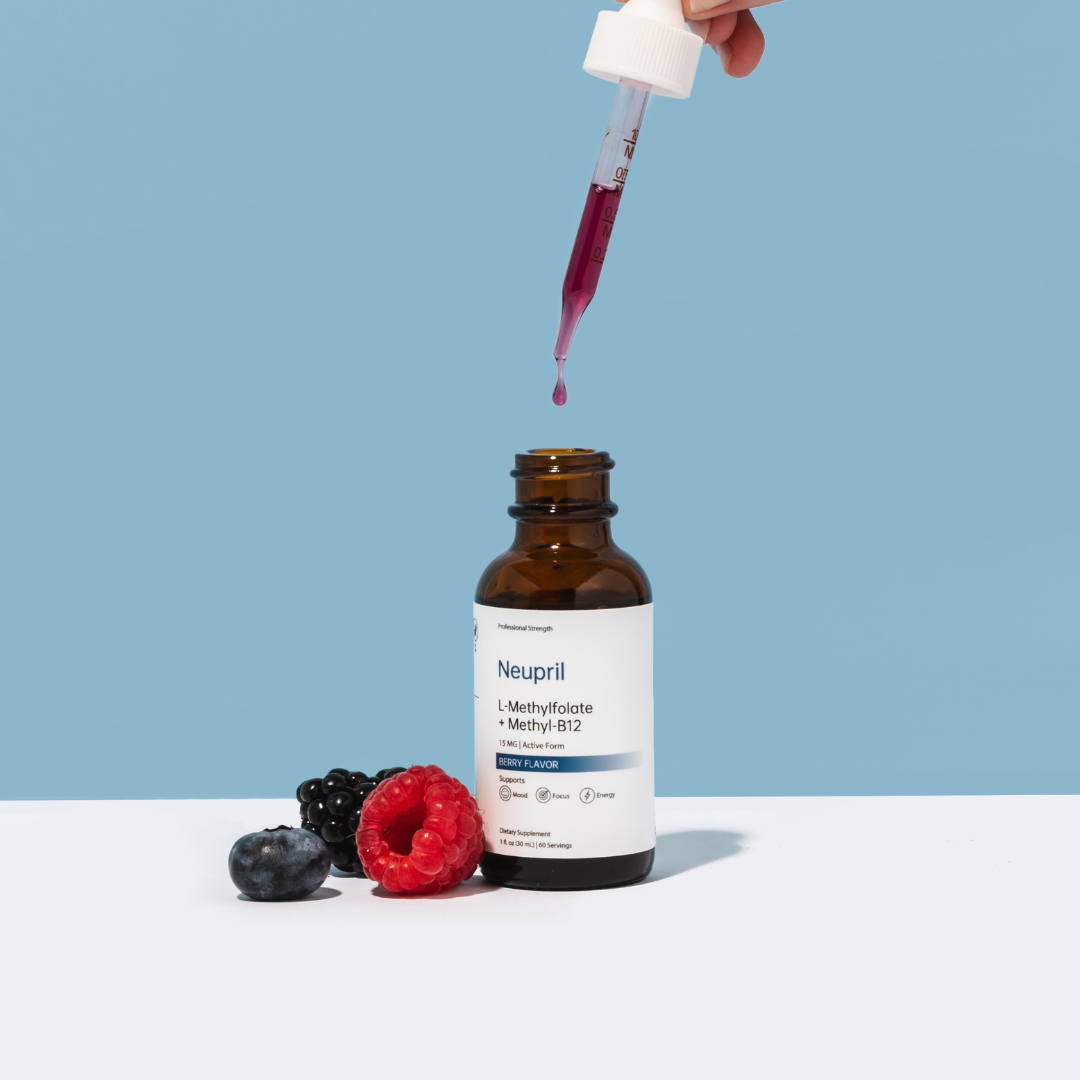 A hand holding a glass dropper dripping pink liquid into an open bottle of Neupril L-Methylfolate + Methyl-B12 supplement, styled on a white surface with a fresh raspberry, blackberry, and blueberry against a blue background.
