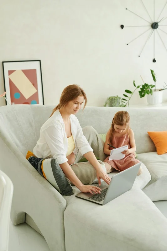 Woman using a laptop on a couch with a child writing on paper.
