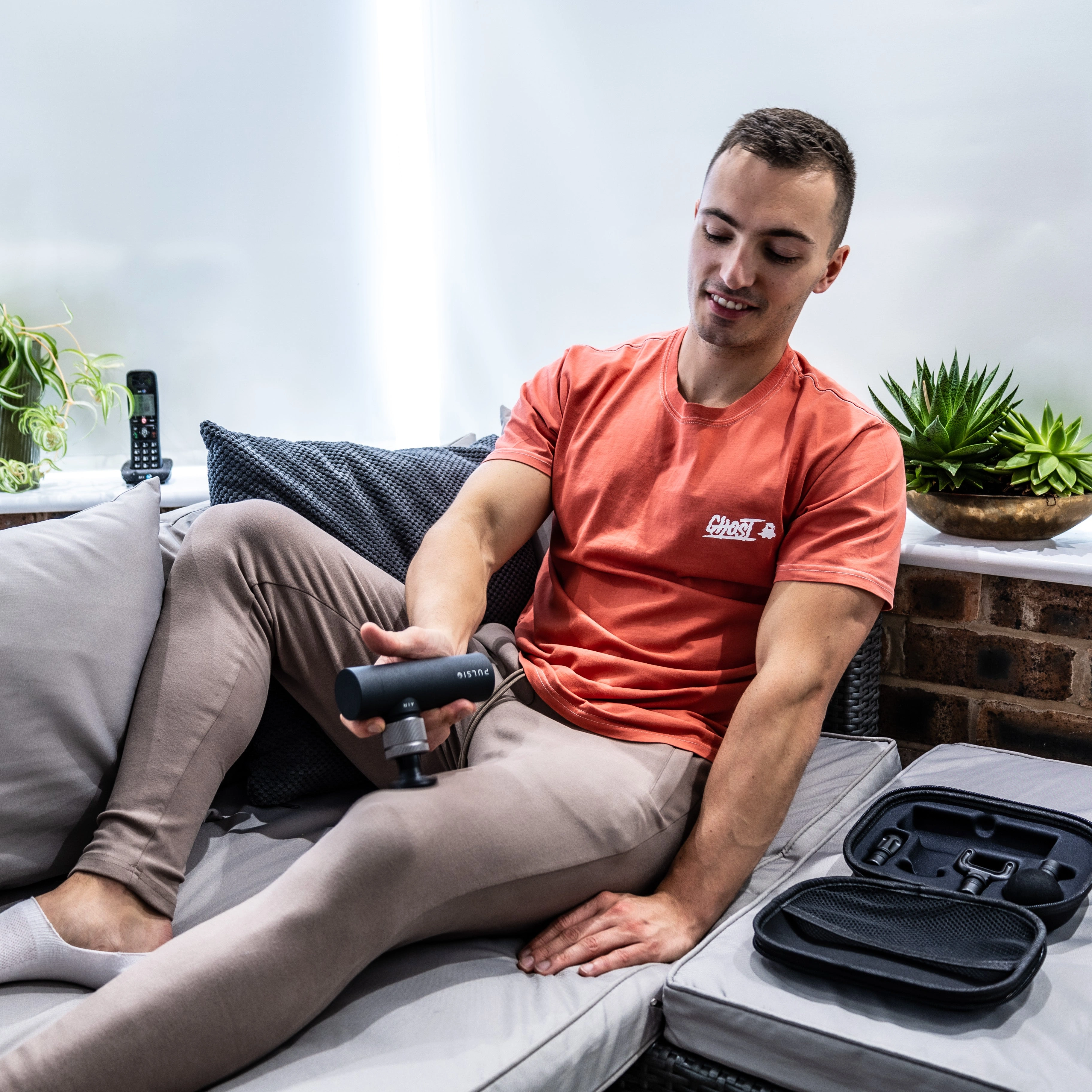 Man using a massage gun on his thigh while sitting on a sofa.