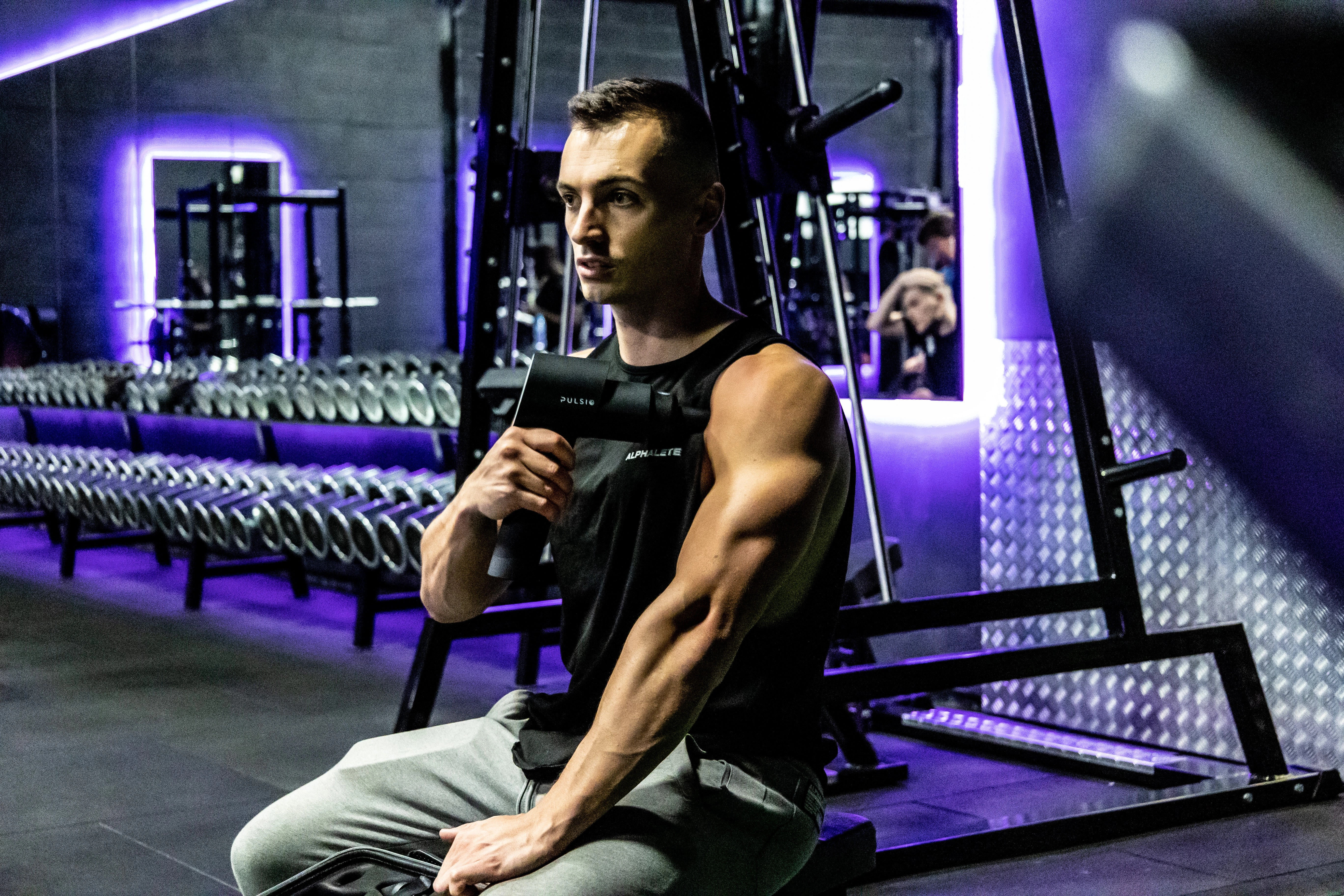 Man holding a massage gun in a gym with weights in background.