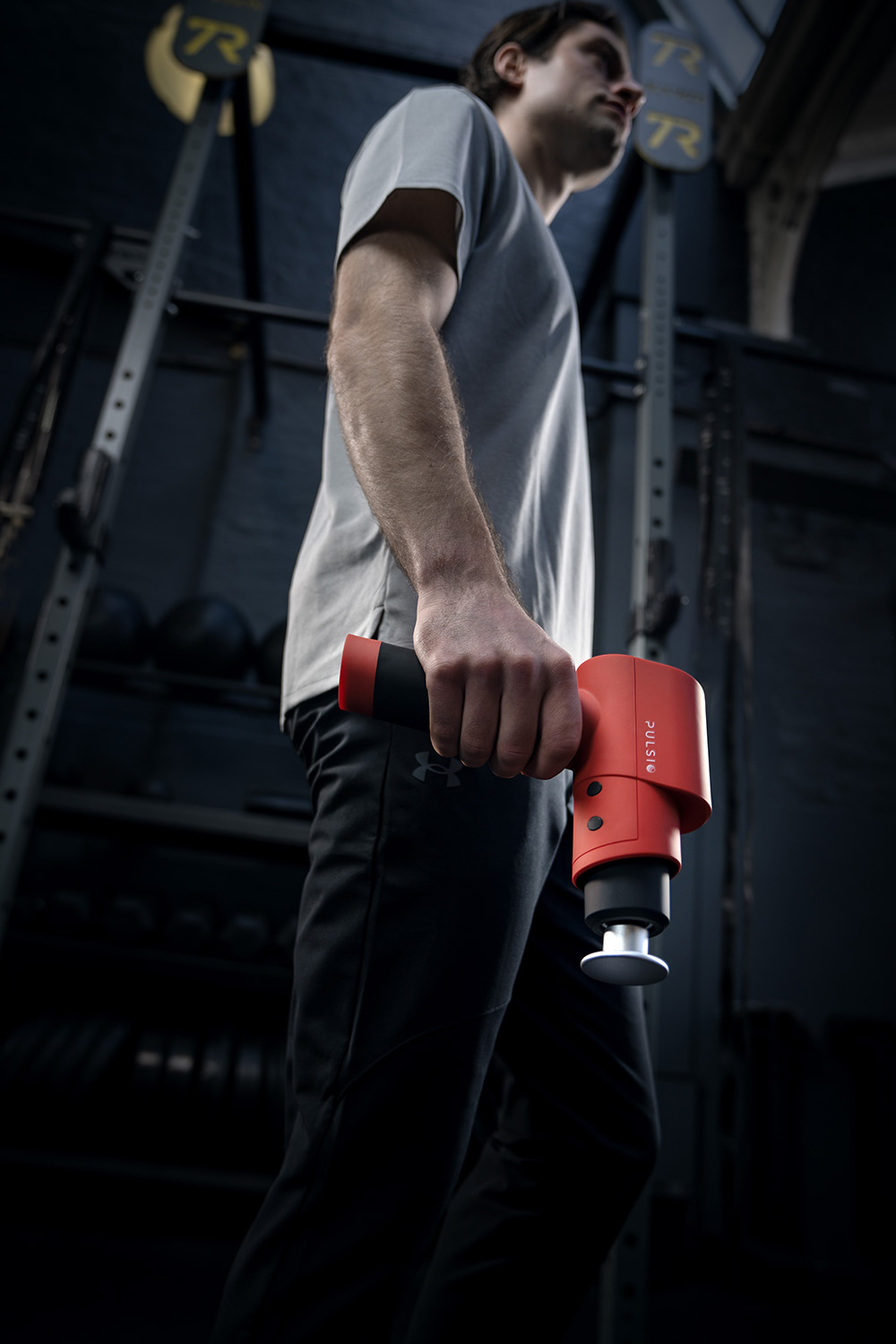 A man in athletic wear standing in a gym while holding a red massage gun.