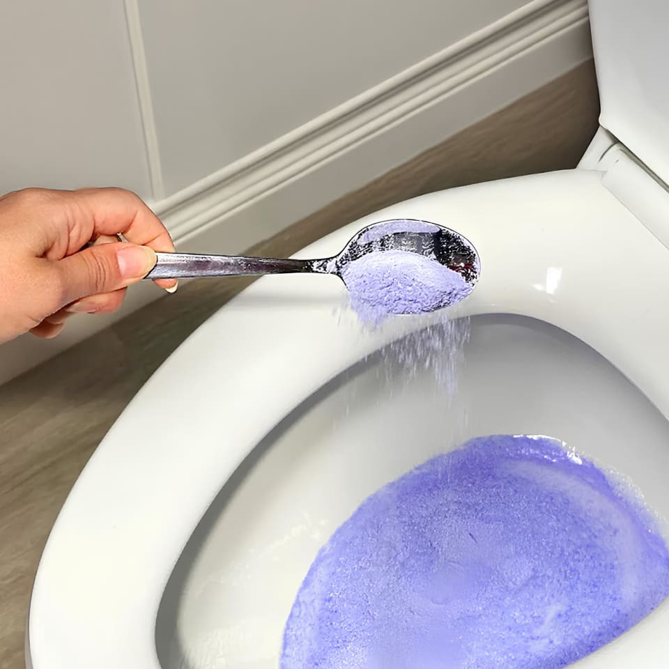 Hand pouring purple powder into a toilet bowl with a spoon.