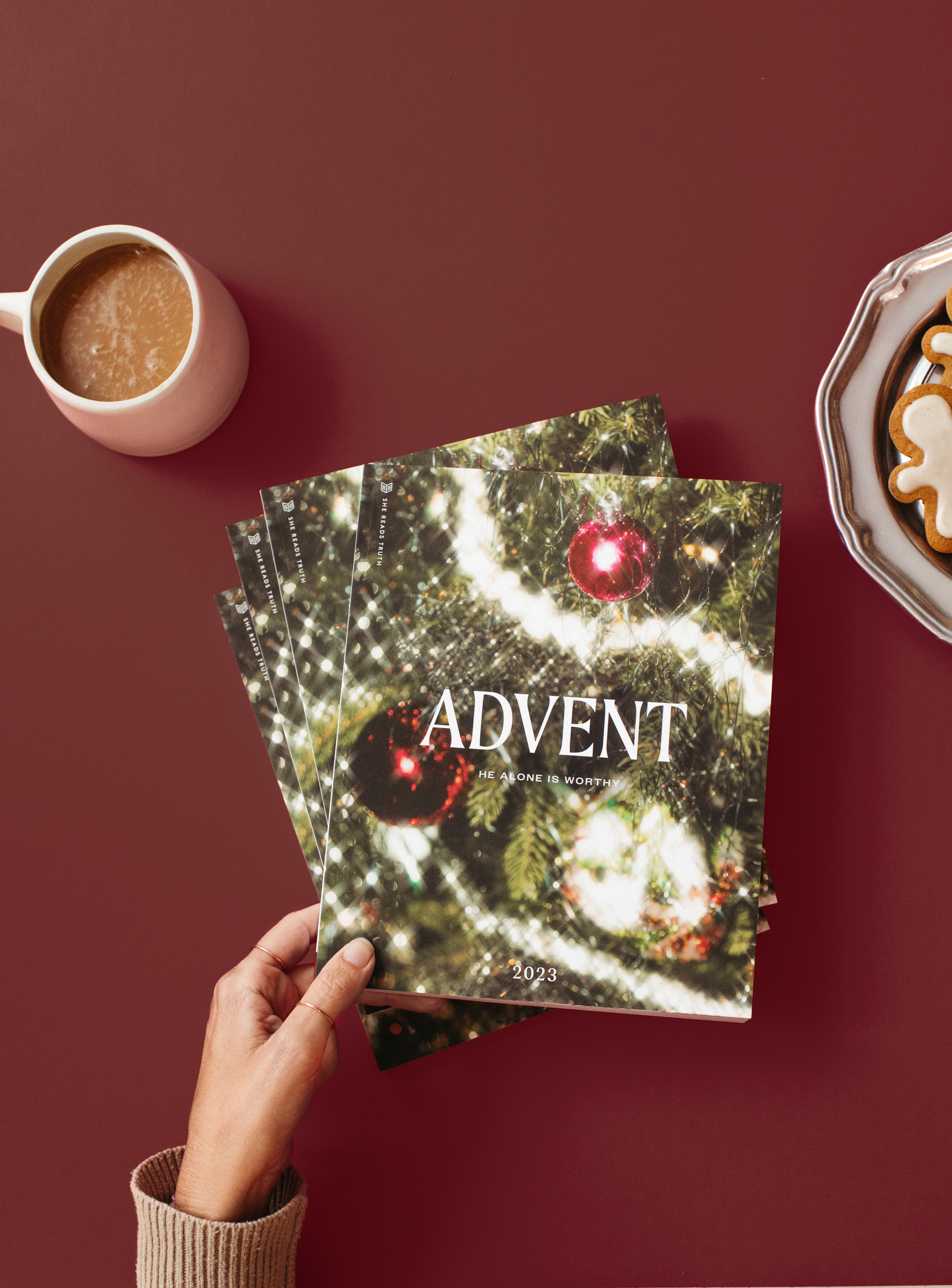 Hand holding Advent booklets, coffee cup, and cookies on a red surface.