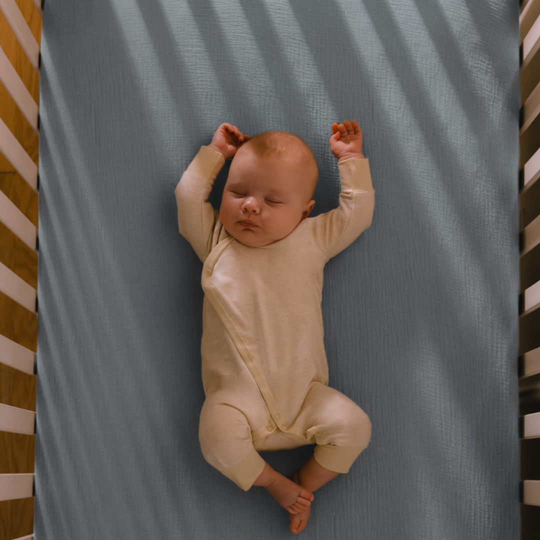 An overhead view of a baby sleeping on its back in a crib on a grey-blue sheet.