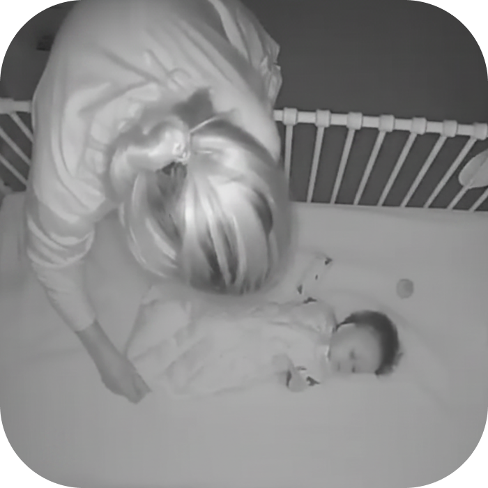 A black and white image of a person with light hair leaning over a baby sleeping in a crib.