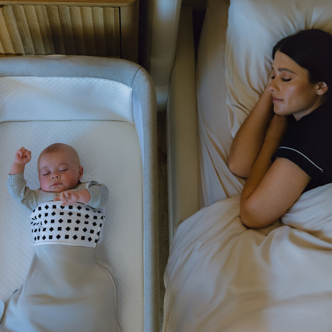 An overhead view shows a baby sleeping in a bassinet next to a woman sleeping in a bed.