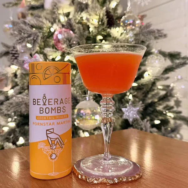 Cocktail glass with orange drink beside a Beverage Bombs container, in front of a decorated Christmas tree.