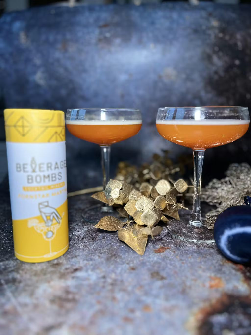 Two orange cocktails in coupe glasses sit next to a 'Beverage Bombs' container and gold decorations.