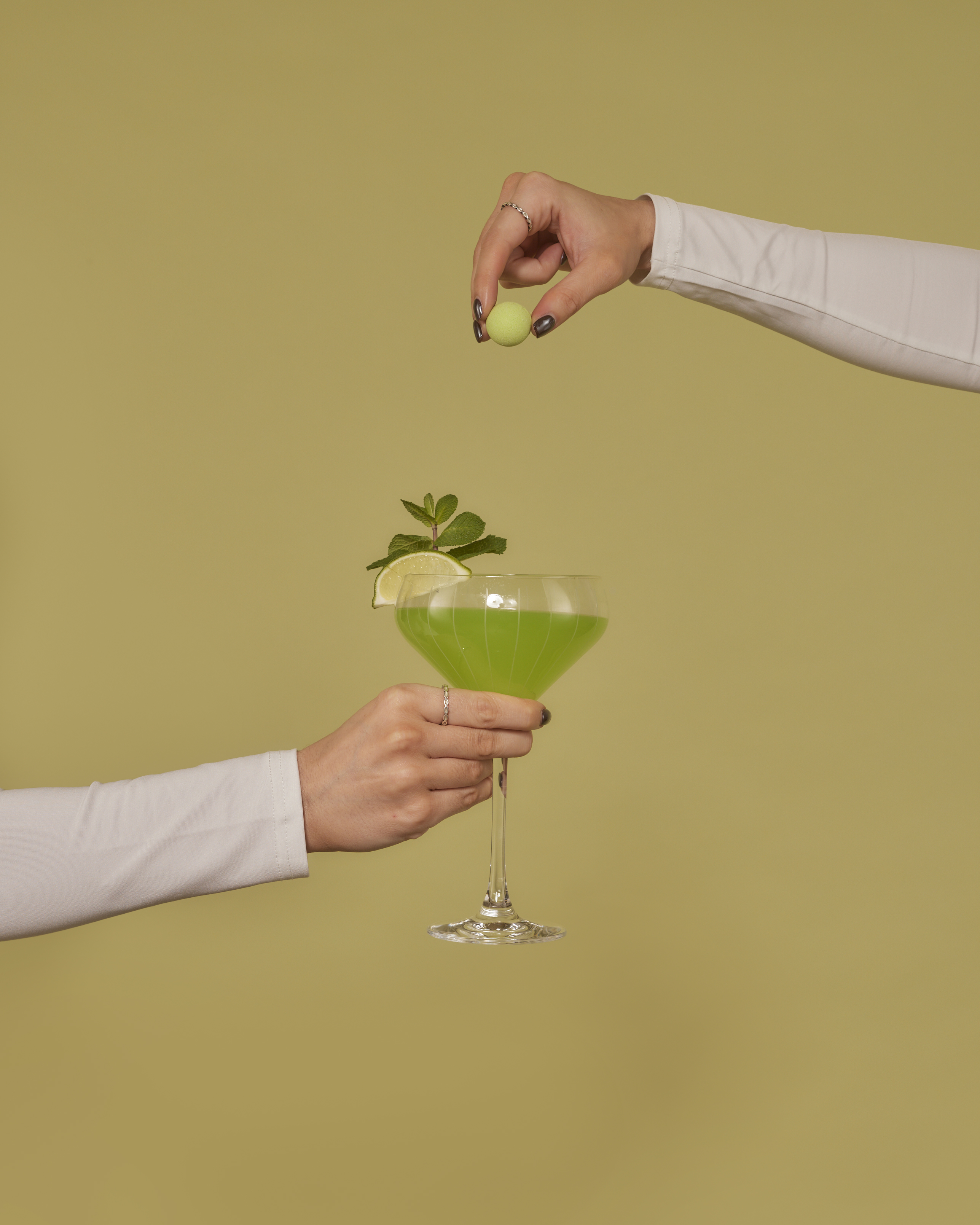 A hand drops a small green ball into a green cocktail held by another hand against a green background.