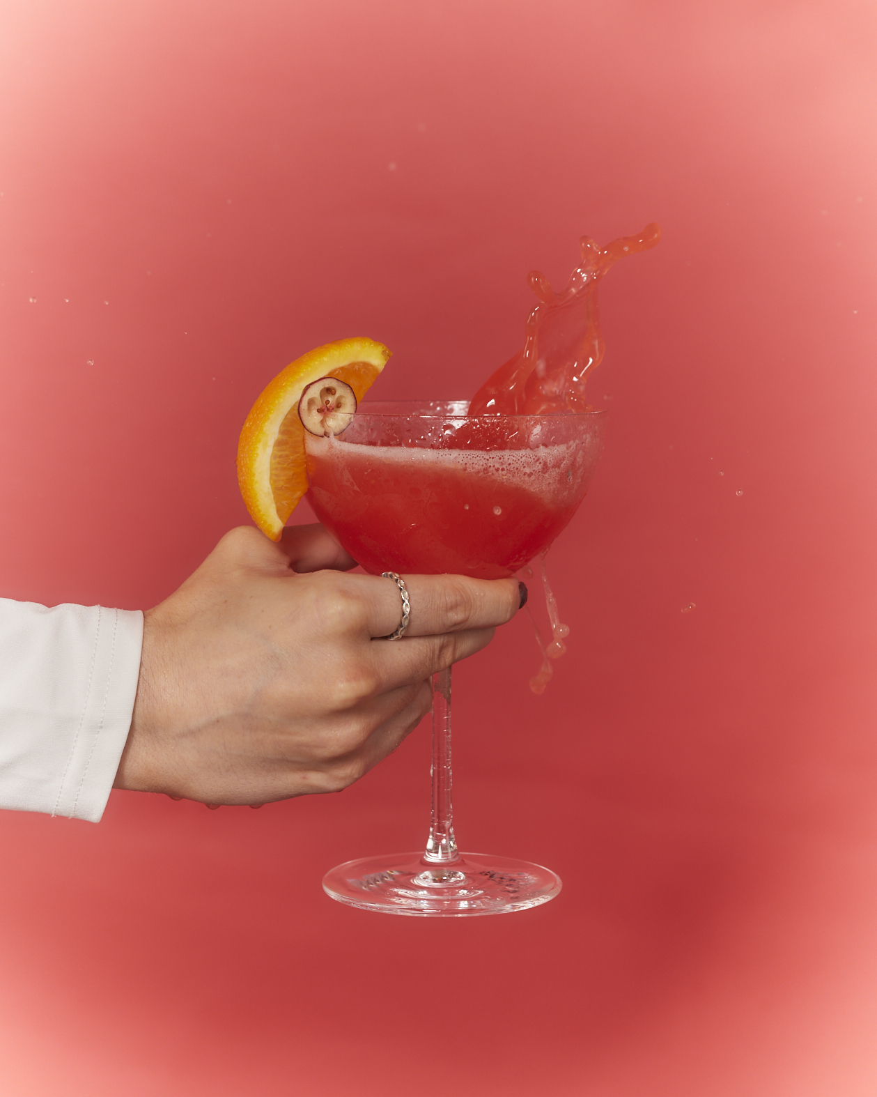A hand holds a red cocktail that is splashing, garnished with an orange slice against a red background.