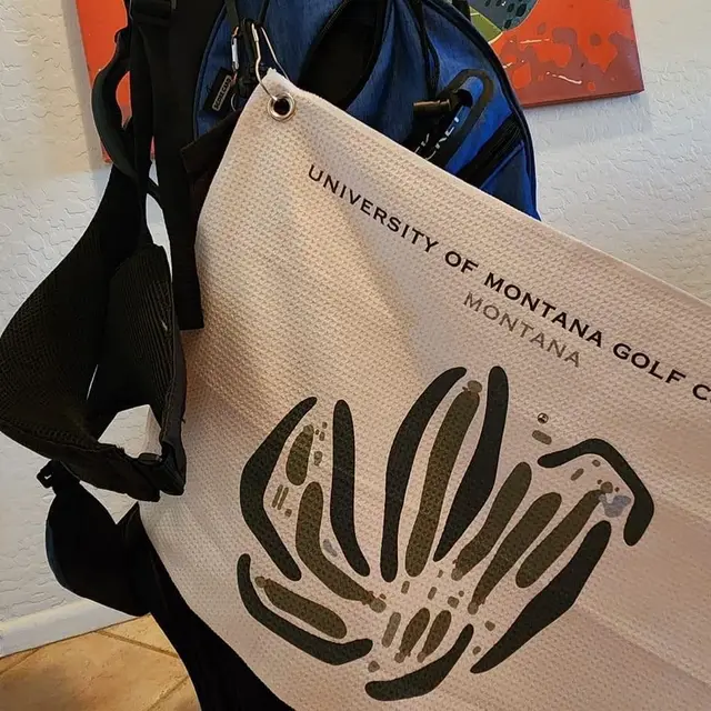 A University of Montana Golf Course towel with a course map design hanging from a blue golf bag.