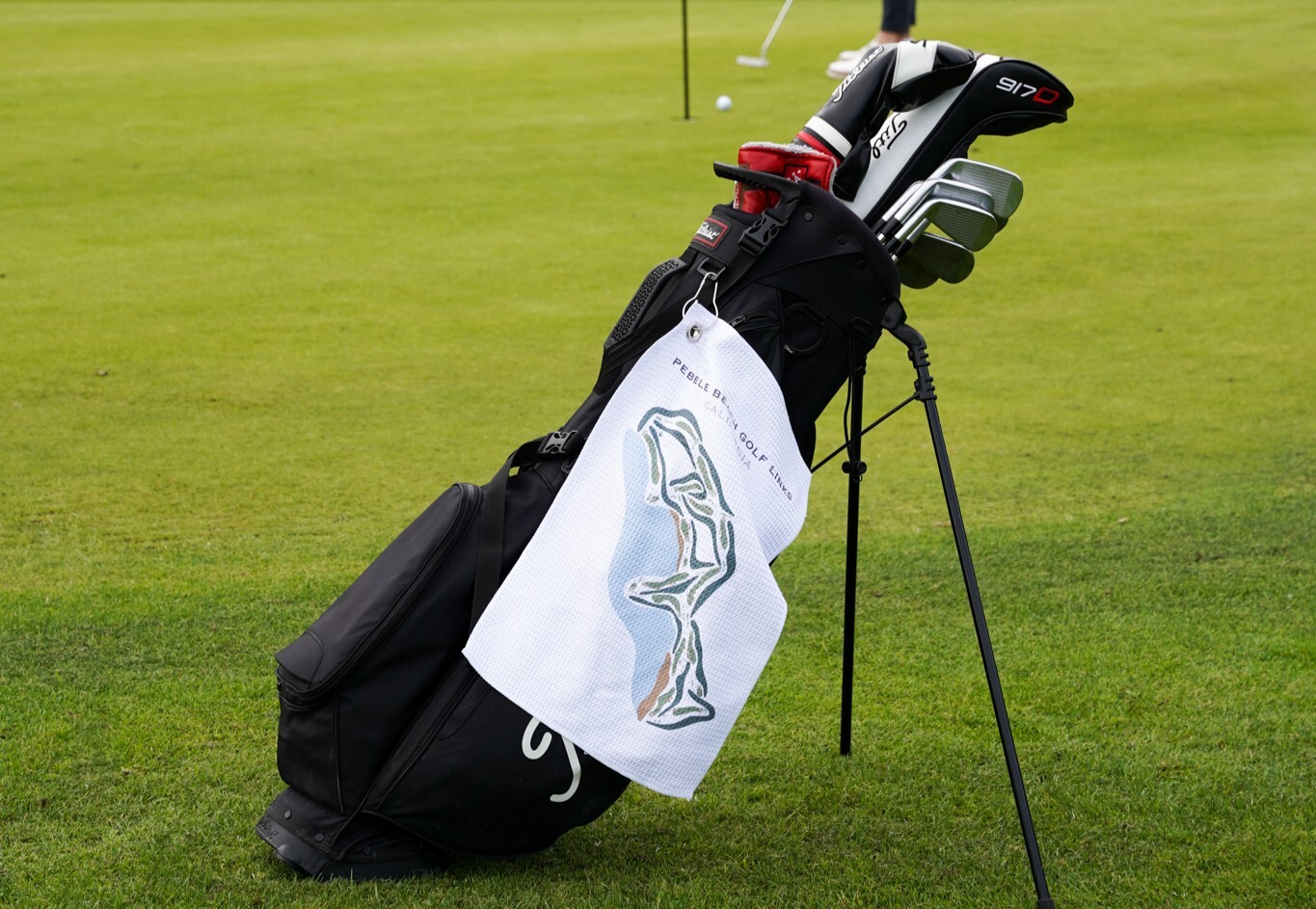 A black golf bag with clubs and a Pebble Beach towel sits on a green golf course.