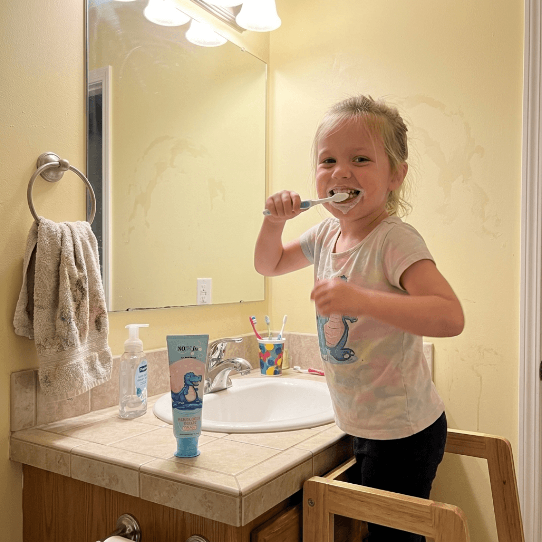 A young girl smiles while brushing her teeth in a bathroom, standing on a wooden stool.