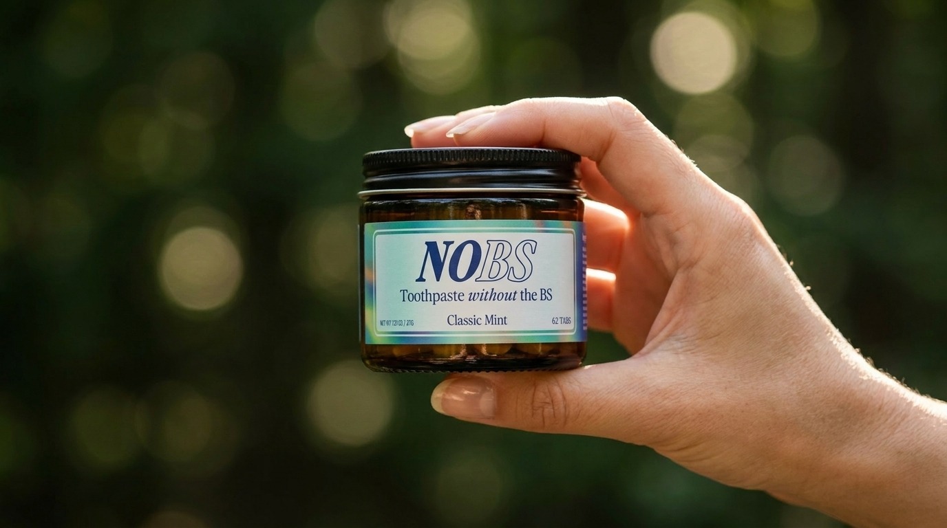 A hand holding a glass jar of NOBS Classic Mint toothpaste tablets against a blurred green background.