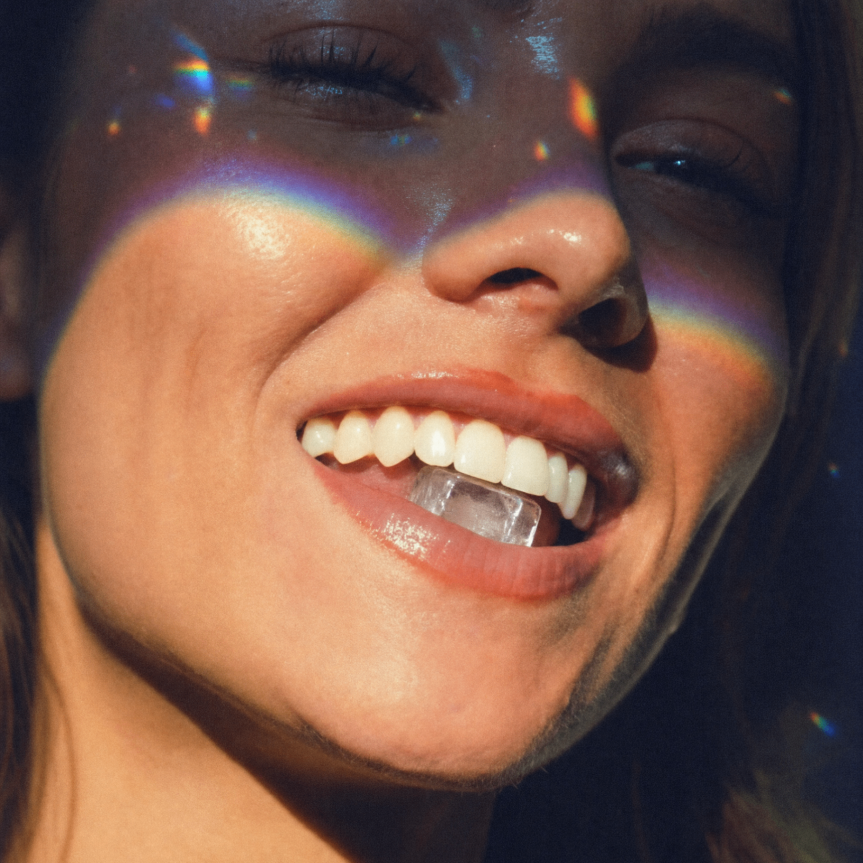 A close-up of a woman smiling with an ice cube in her mouth, with rainbow light on her face.