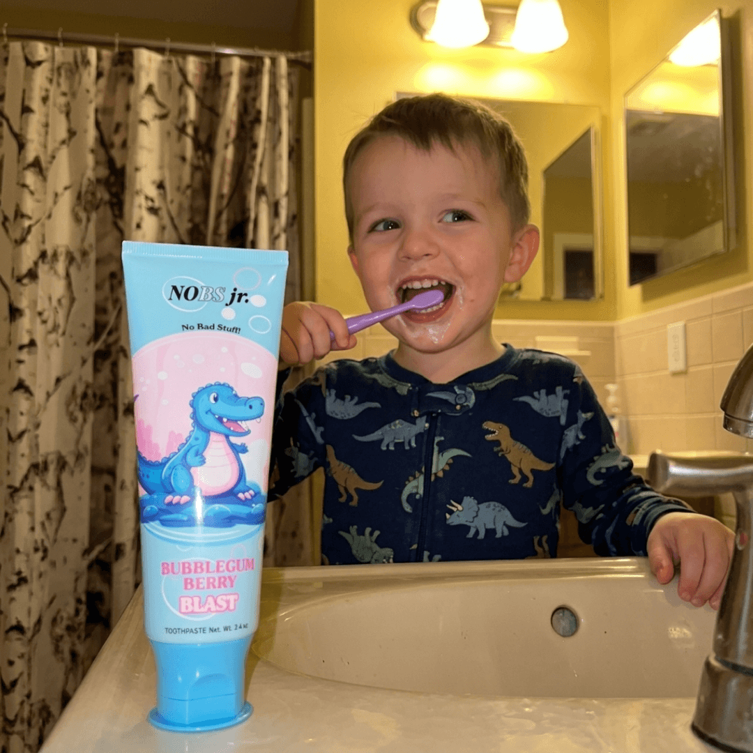 A young boy in dinosaur pajamas happily brushes his teeth at a bathroom sink next to a toothpaste tube.