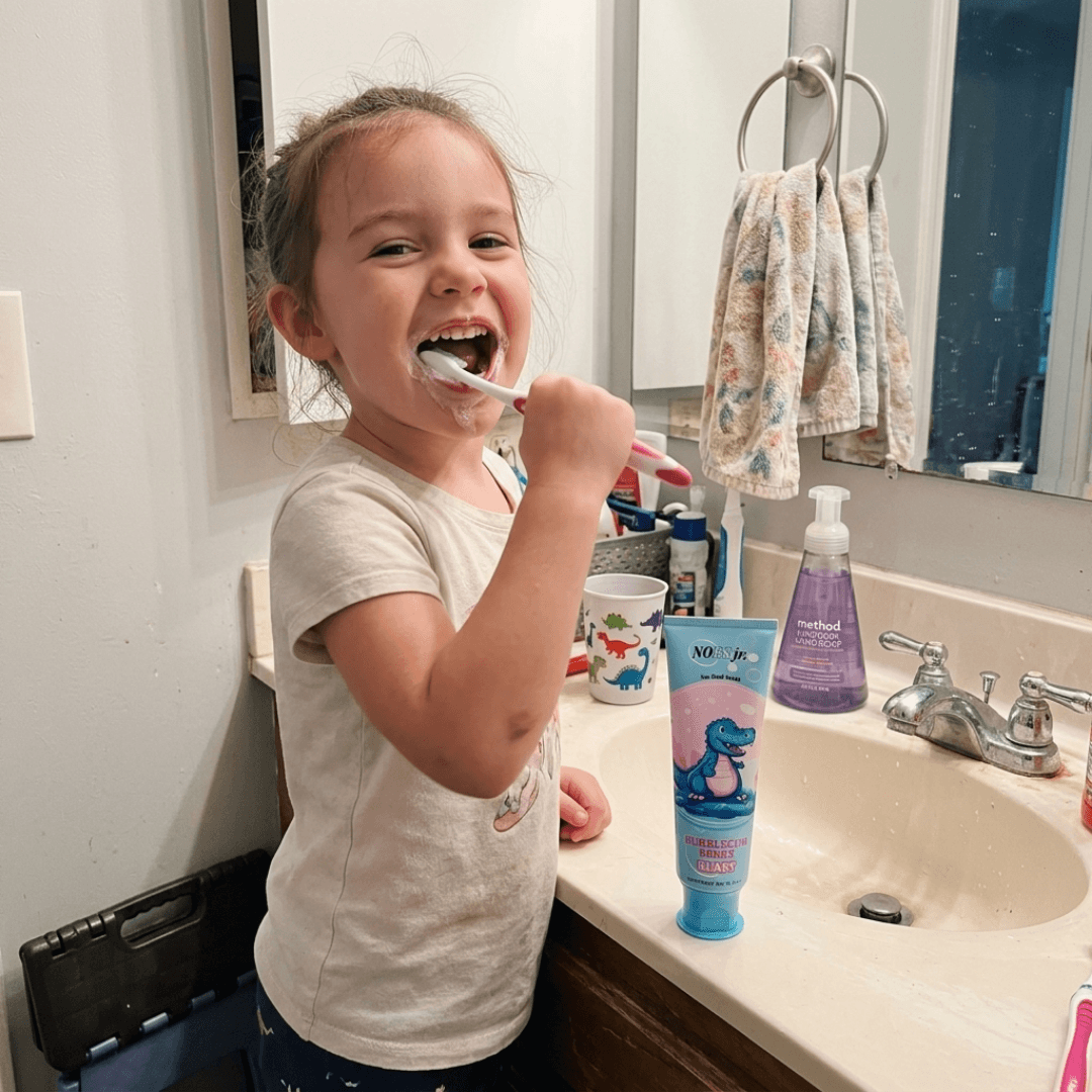 A young girl with her hair up smiles while brushing her teeth in a bathroom.