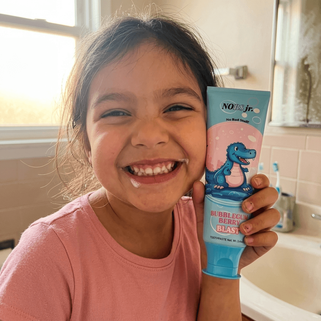A smiling young girl with toothpaste foam on her face holds up a tube of toothpaste.
