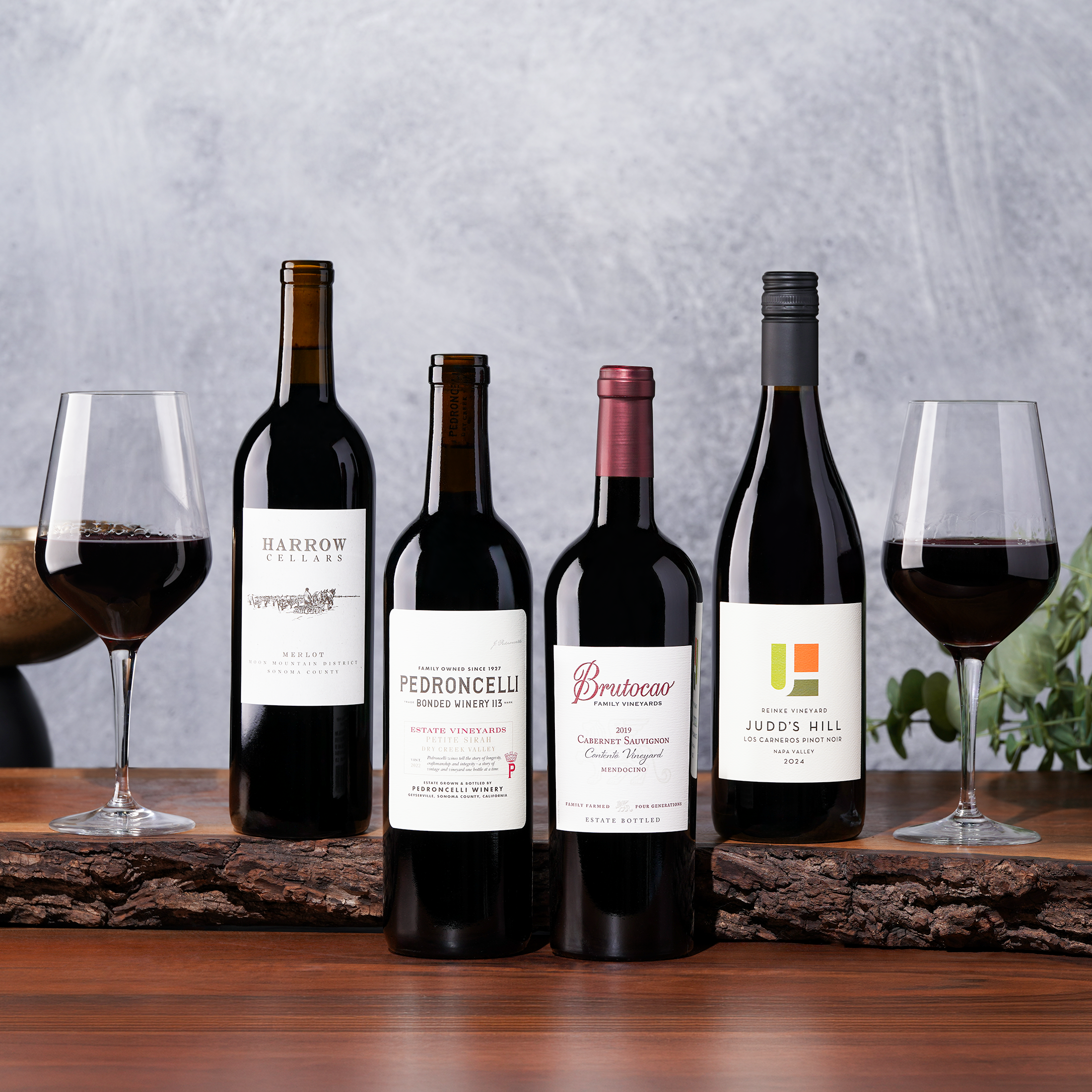 Four bottles of red wine and two full glasses are displayed on a wooden slab.