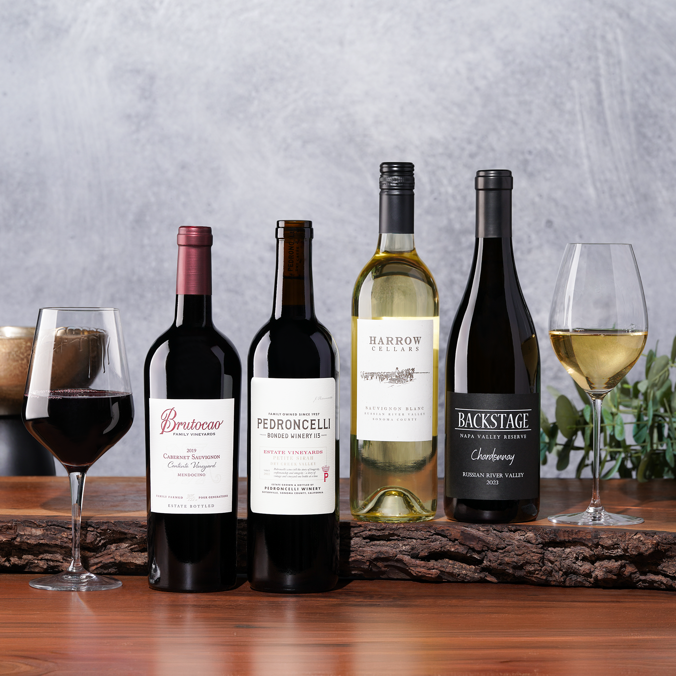 Four bottles of red and white wine with two filled glasses on a rustic wooden plank.