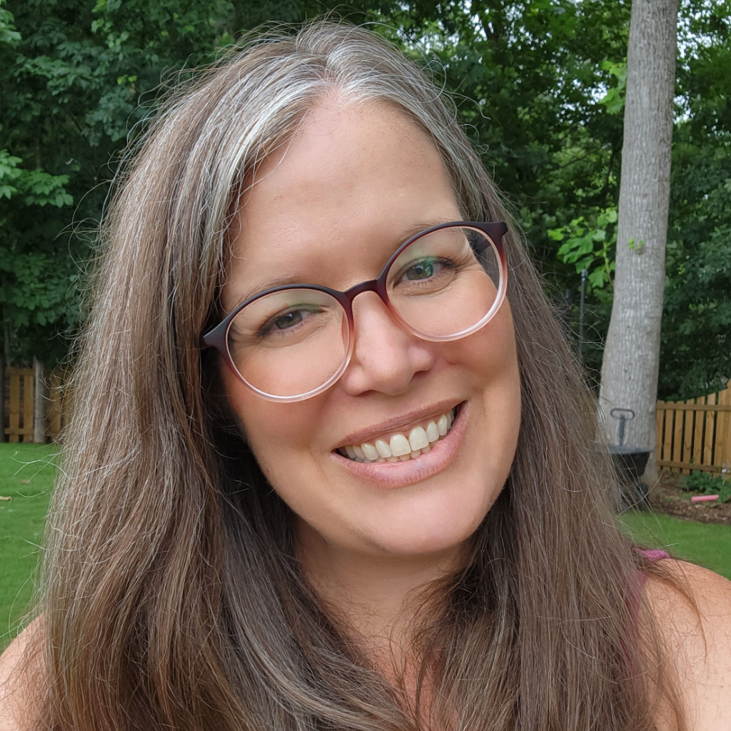 Smiling person with glasses and long hair, outdoors with trees in the background.