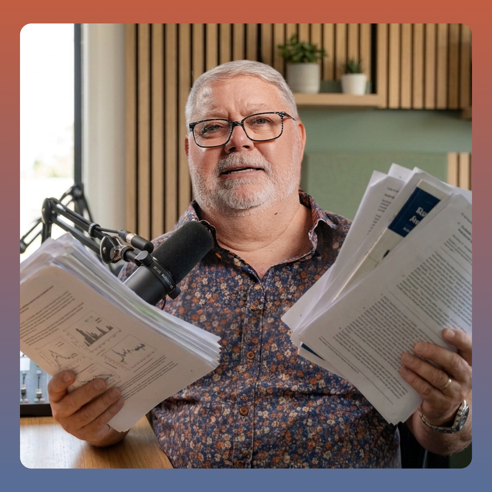 A man with glasses sits at a desk with a microphone, holding up two large stacks of papers.