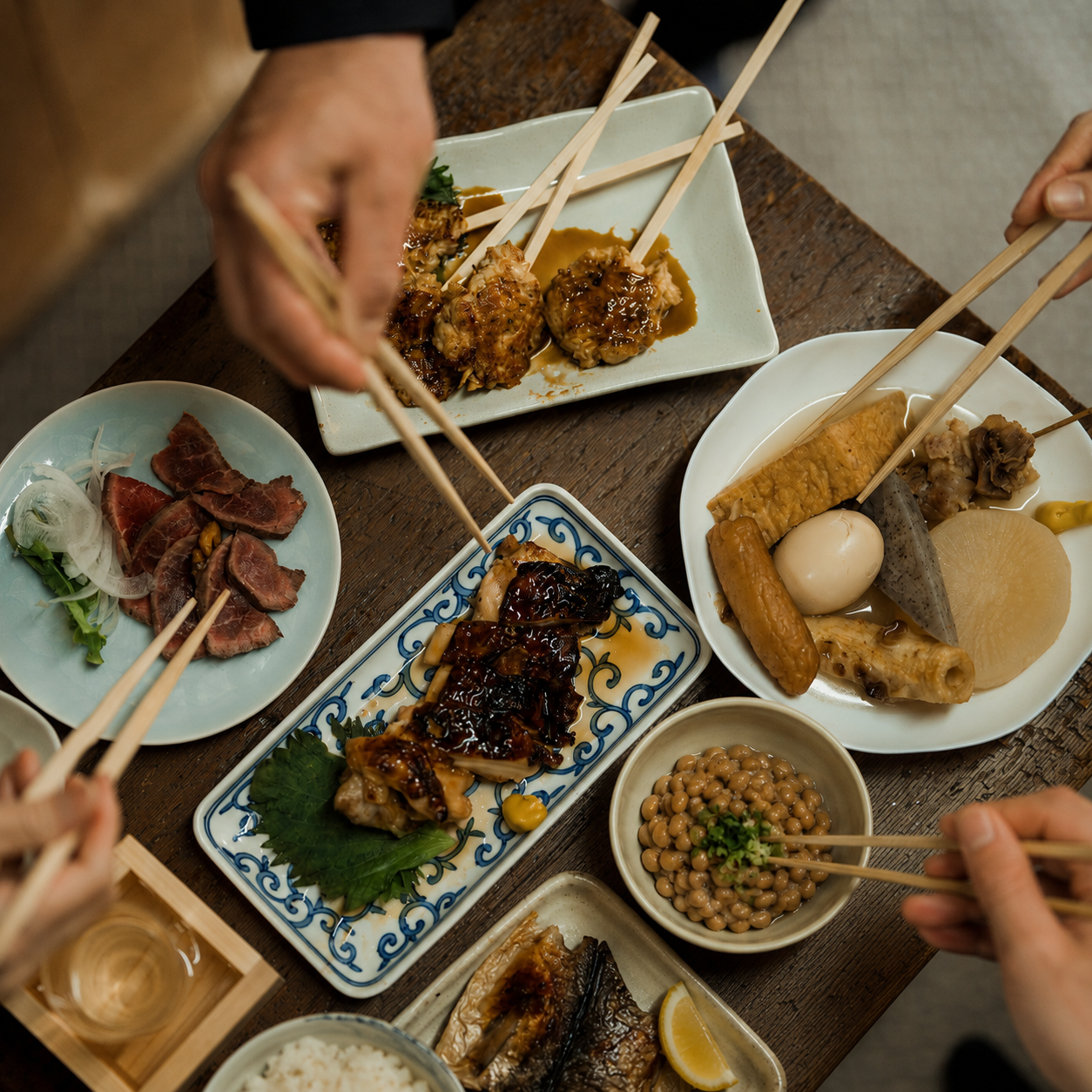 An overhead view of people using chopsticks to eat a variety of Japanese dishes from shared plates.