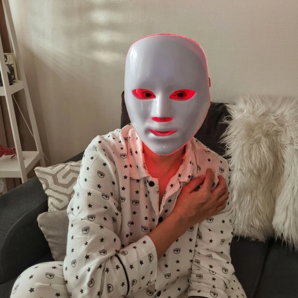 A person in pajamas sits on a couch wearing a white facial mask with glowing red lights.