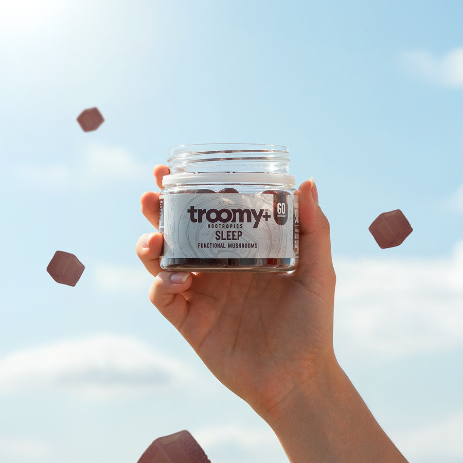 A hand holds a jar of Troomy+ Sleep gummies against a blue sky as several gummies float around.
