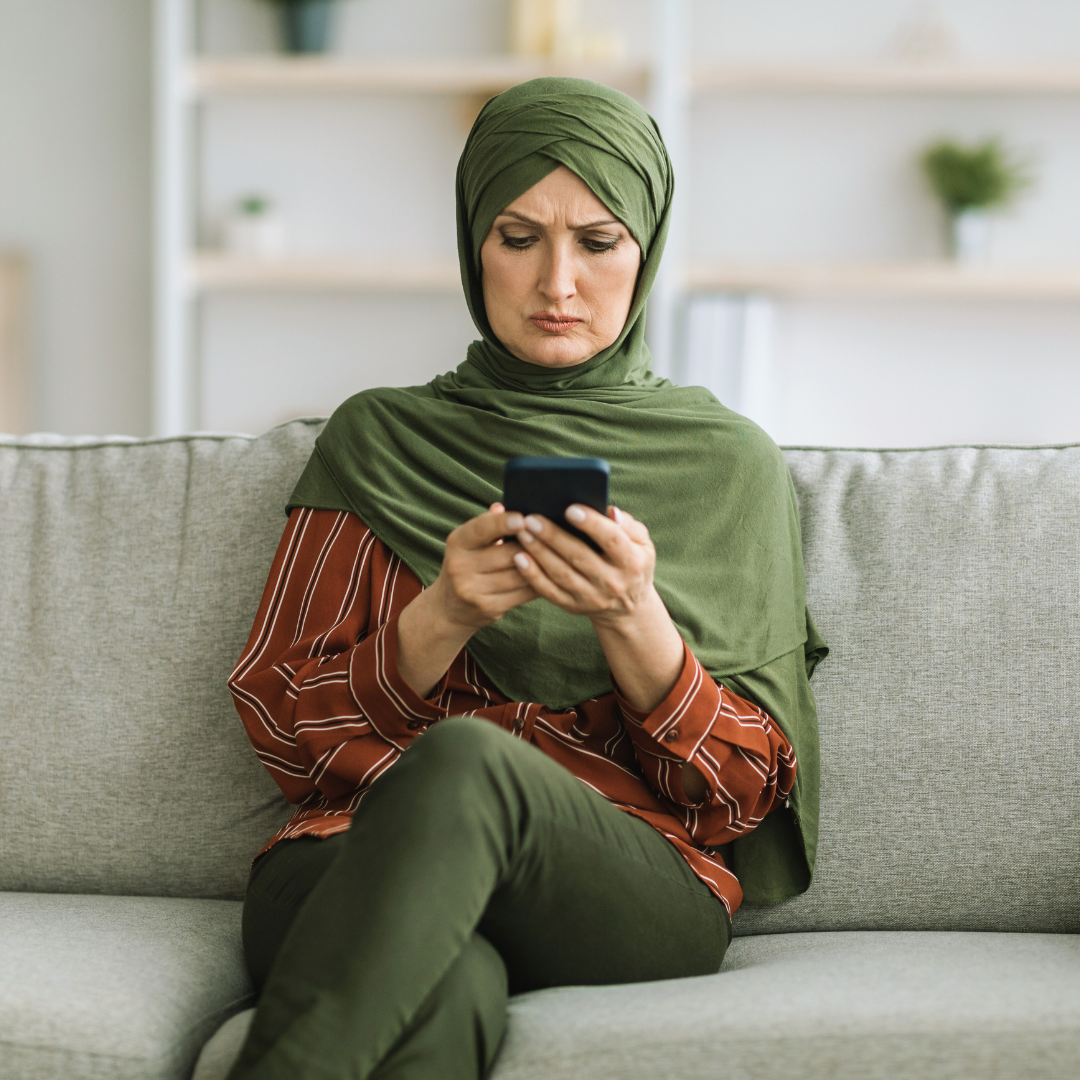 A woman in a green hijab sits on a couch and looks at her smartphone with a concerned expression.