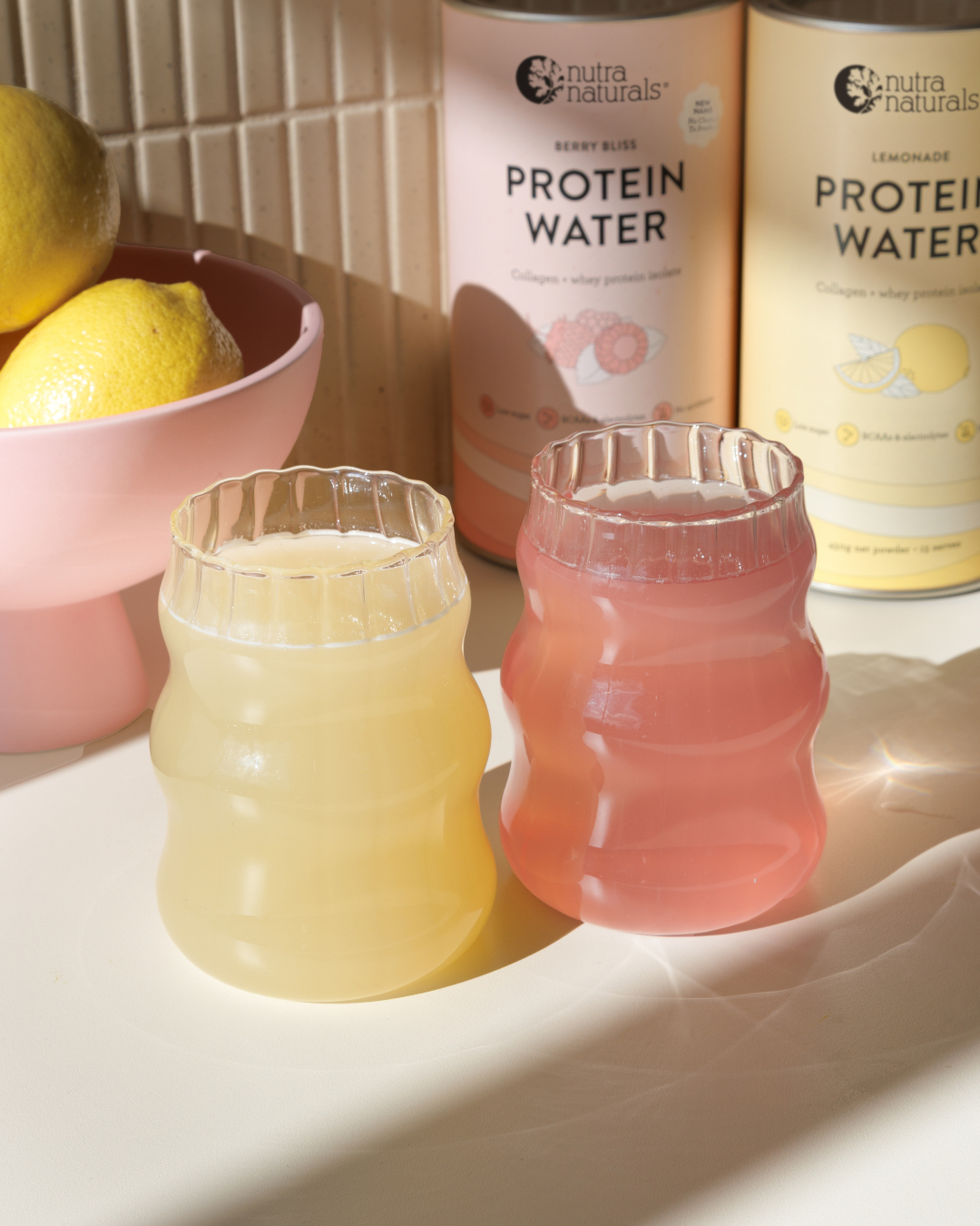Two glasses of yellow and pink protein water sit in front of their containers and a bowl of lemons.