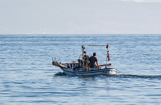 Two people in a small white fishing boat on the water with a hazy shoreline in the background.