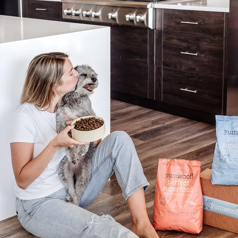 Person kissing small dog, holding bowl, with dog food bags nearby in kitchen.