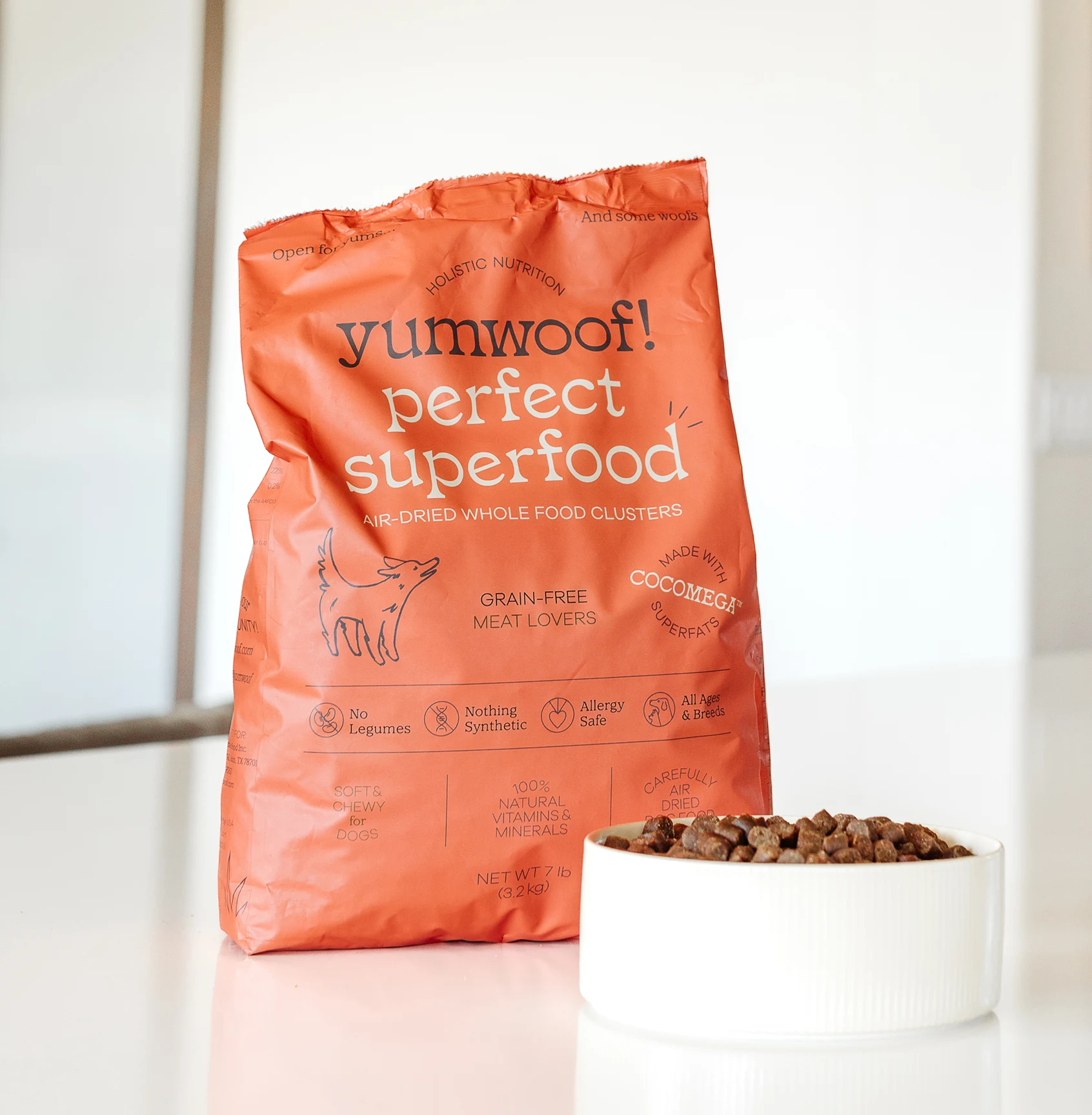 A bag of Yumwoof Perfect Superfood dog food sits on a white table next to a full bowl.